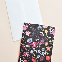 1: Notecard with colorful floral decoration on black background, and the text Congratulations in metallic pink script, with white envelope.
