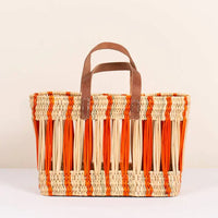 Small / Clementine: An oblong open weave basket in natural reed with bright orange stripes and brown leather handles.