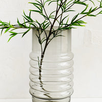 2: A tall grey glass vase with curved ribbing at middle, holding leafy branch.