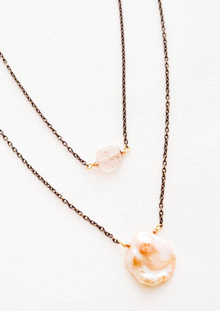 Close up of two layer necklace composed of one dark gold chain with translucent pink crystal and one of dark gold chain and pink freshwater pearl.