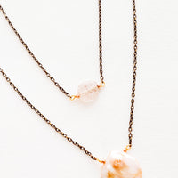 1: Close up of two layer necklace composed of one dark gold chain with translucent pink crystal and one of dark gold chain and pink freshwater pearl.