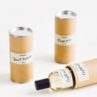 2: Three cardboard cylinder containers with white labels and black text. One tipped on side with glass perfume bottle inside.