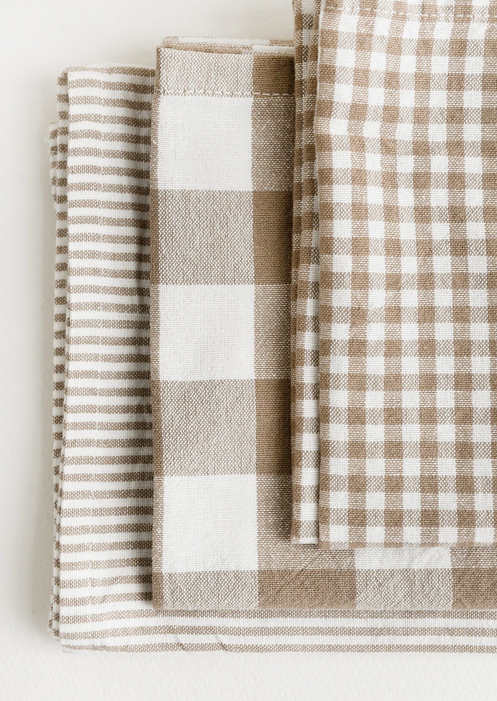 2: Three folded tea towels in tan and white check and stripe patterns.