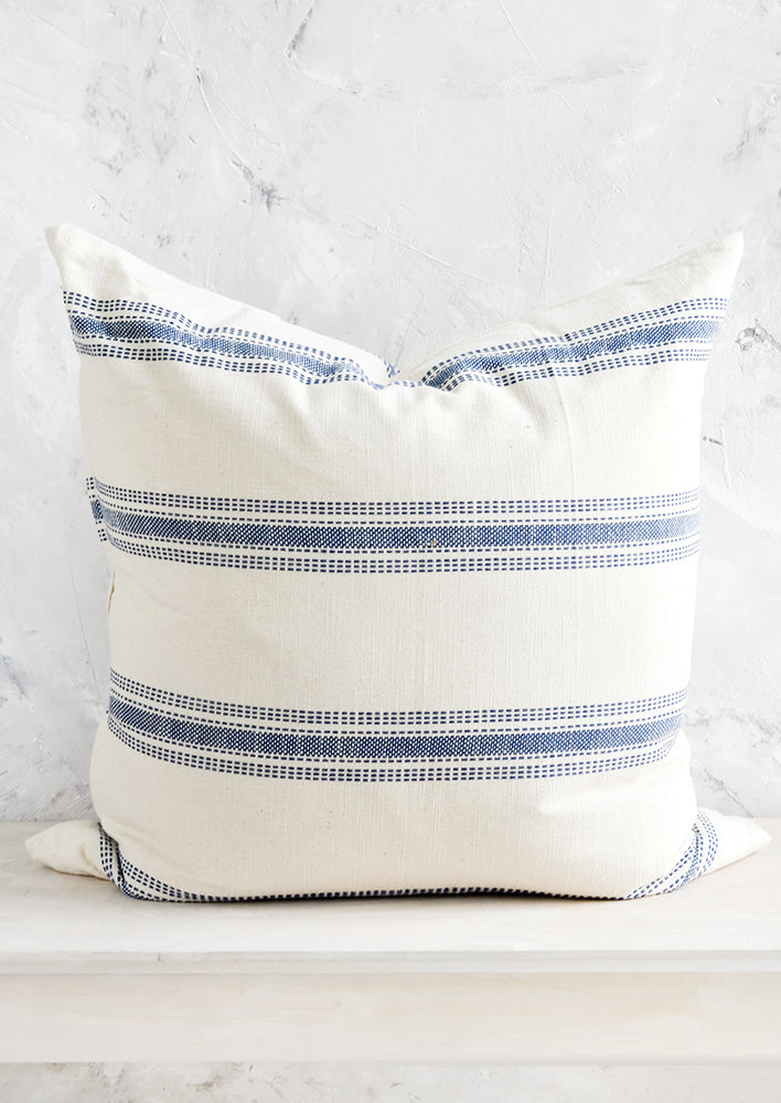 A large, square-shaped throw pillow in cream cotton with horizontal embroidered blue stripes.