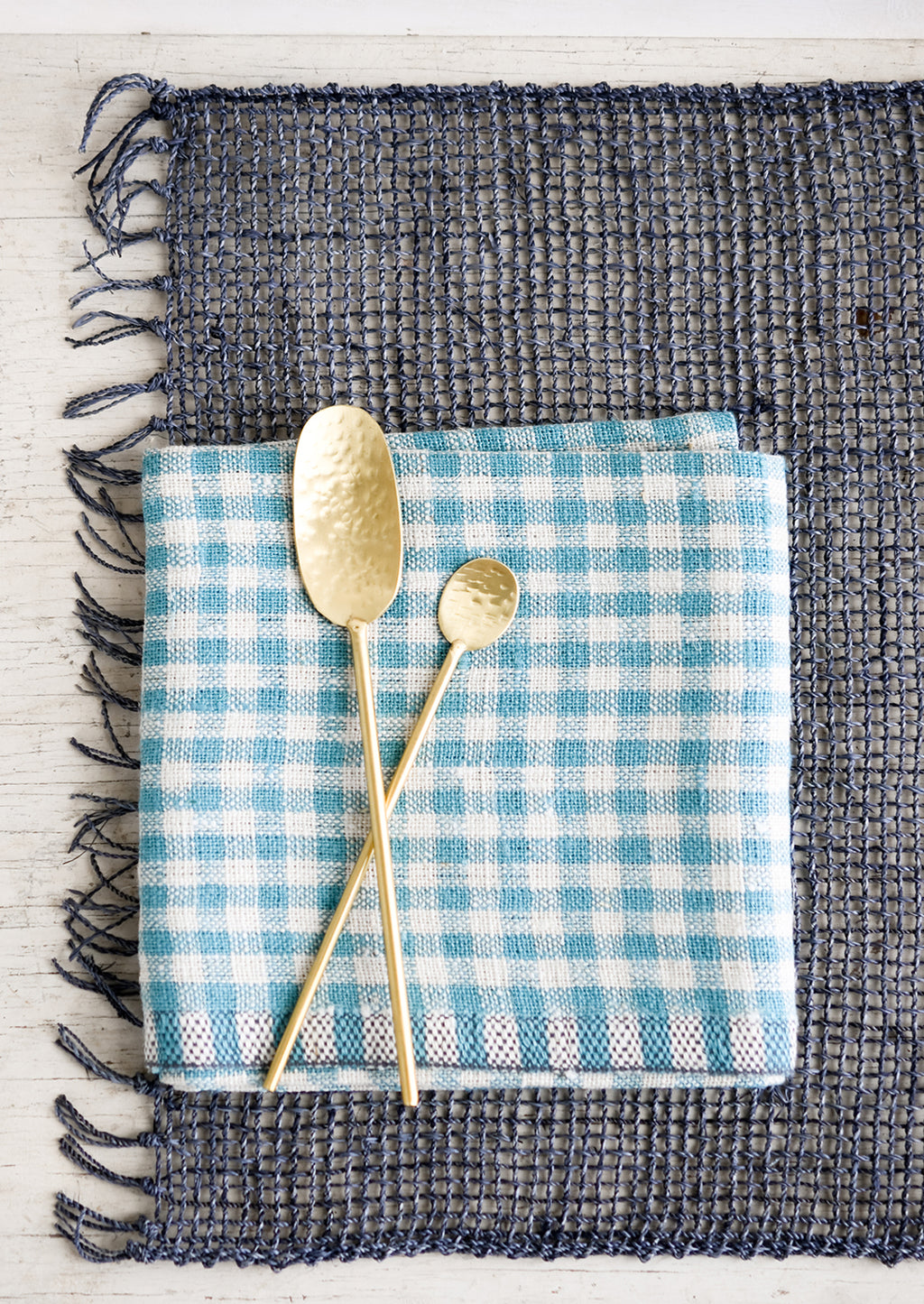 1: A blue-grey woven straw placemat with gingham napkin and gold spoons.