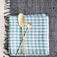 1: A blue-grey woven straw placemat with gingham napkin and gold spoons.