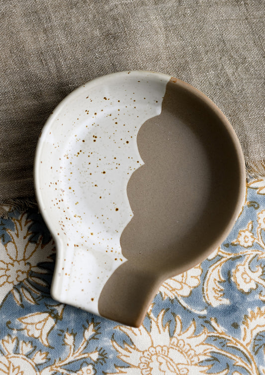 A ceramic spoon rest with scalloped edge dividing brown and speckled white halves.