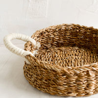 1: A round, shallow tray made from seagrass with ivory jute handles at sides.