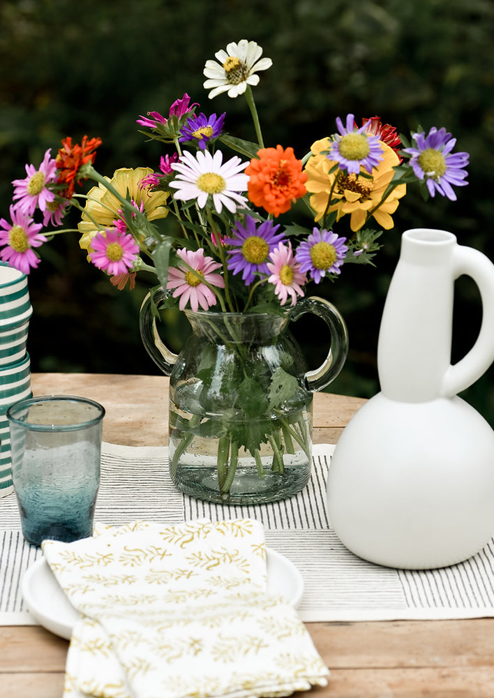 A clear, jug-shaped glass vase on a set table with flowers.