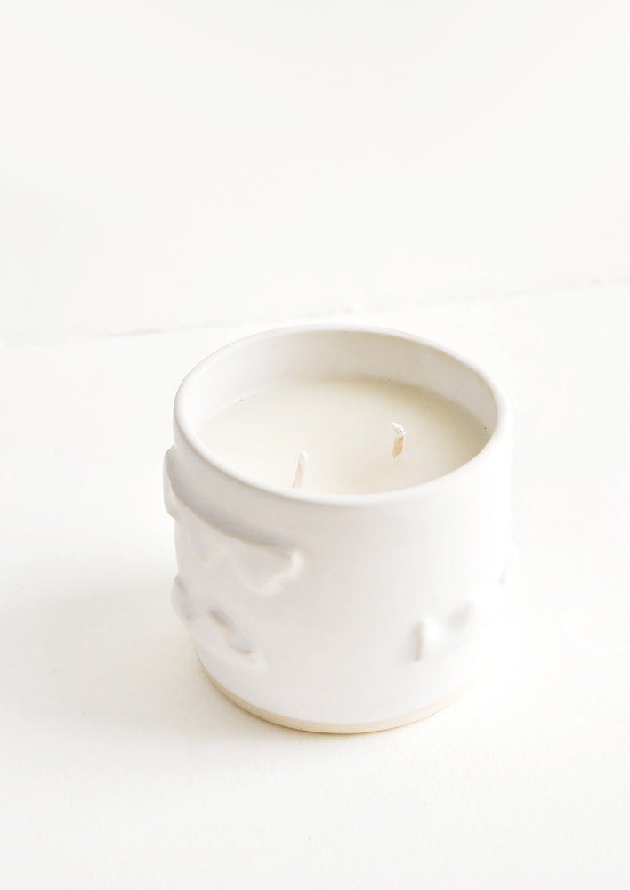 A candle in a white ceramic container affixed with three dimensional squiggles.