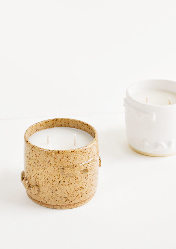A candle in a light brown ceramic container with one in a white ceramic container just behind.