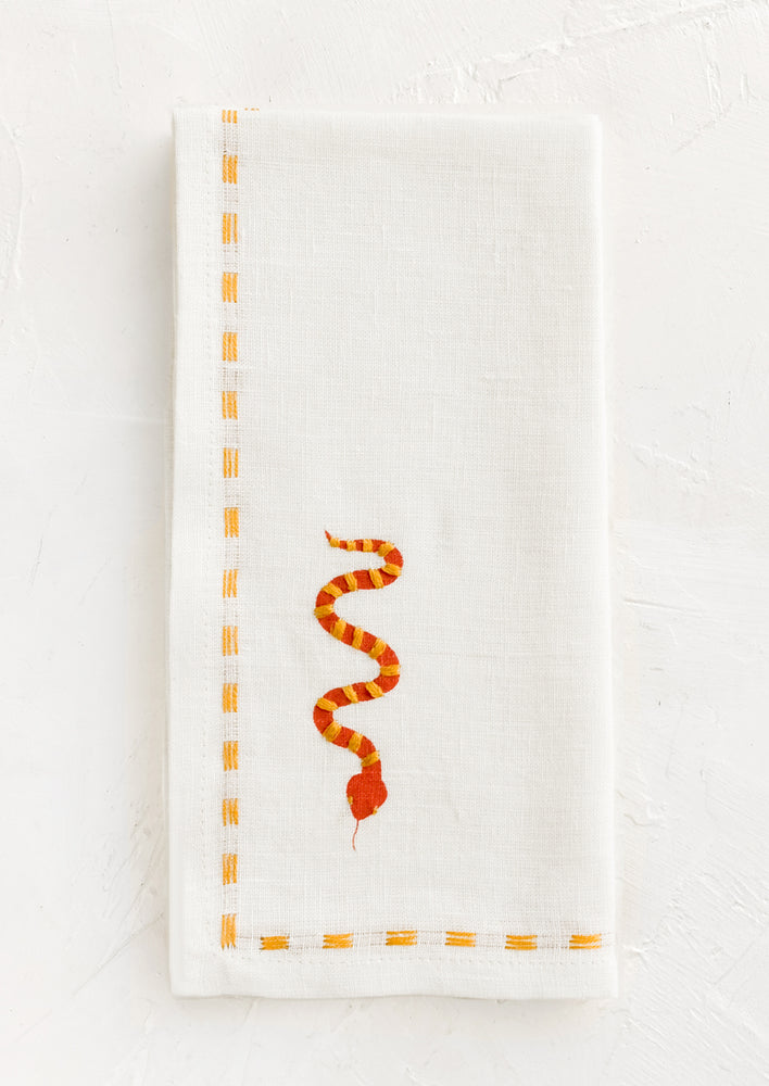 1: A white linen napkin with yellow stitching and red and yellow snake embroidery at corner.