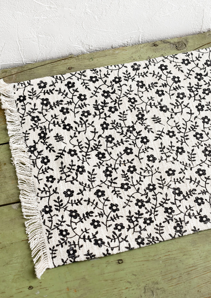 1: A black and white floral print placemat with fringe trim.