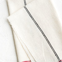 2: A natural cotton napkin with black and red stripe detailing and raw edges.