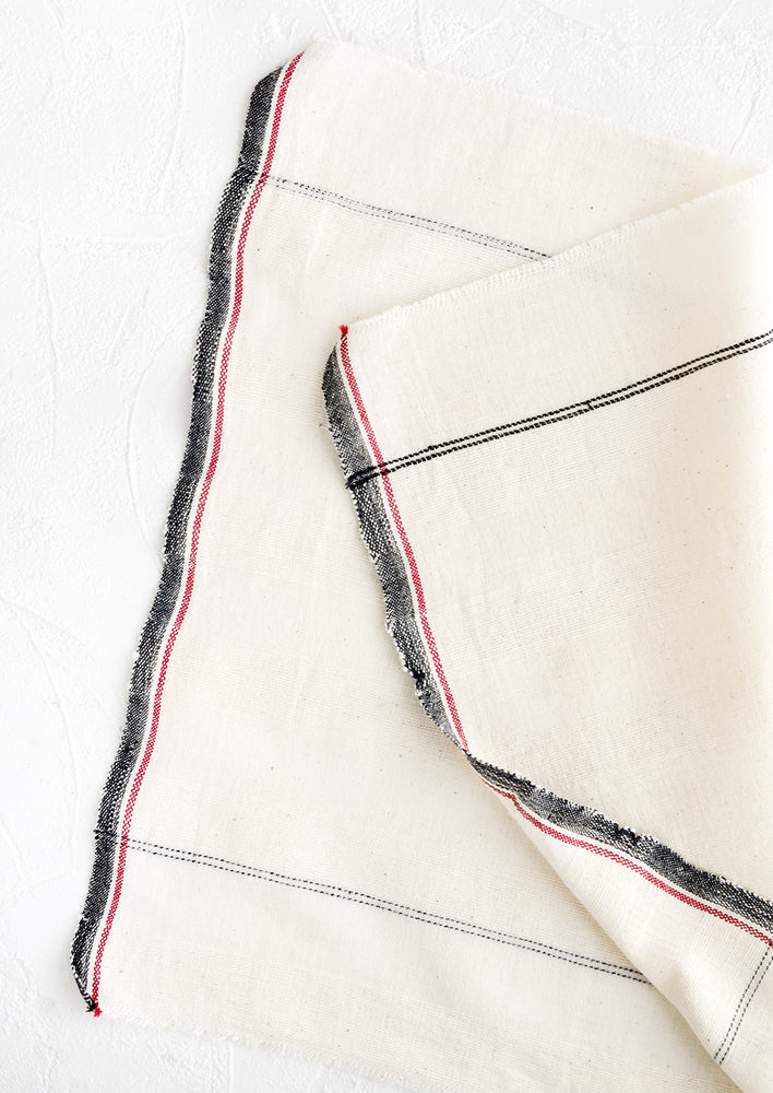 A natural cotton napkin with black and red stripe detailing and raw edges.