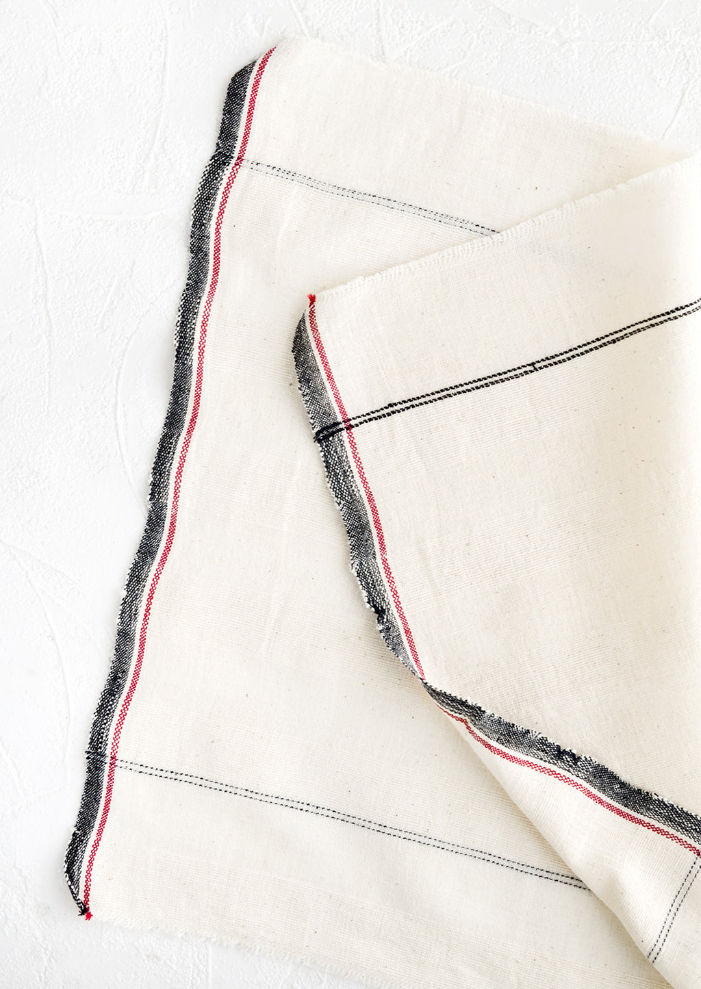 1: A natural cotton napkin with black and red stripe detailing and raw edges.