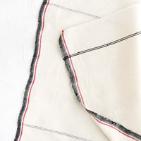 1: A natural cotton napkin with black and red stripe detailing and raw edges.