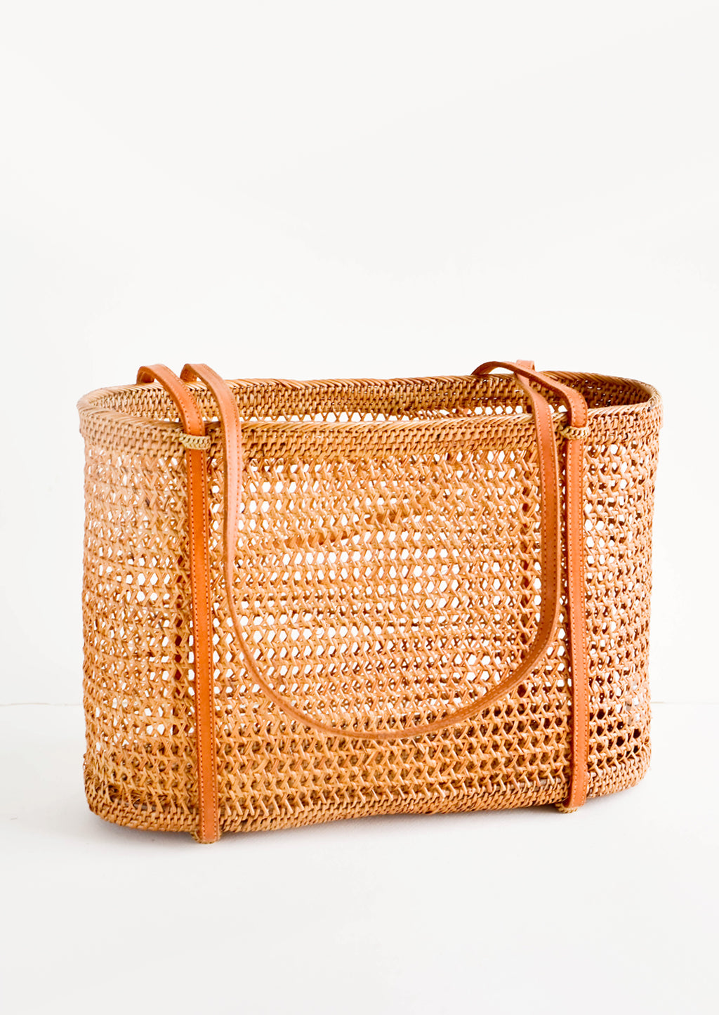 1: East-west tote bag made from open weave rattan, thin, tanned leather shoulder straps wrap around the bottom of the bag