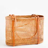 1: East-west tote bag made from open weave rattan, thin, tanned leather shoulder straps wrap around the bottom of the bag