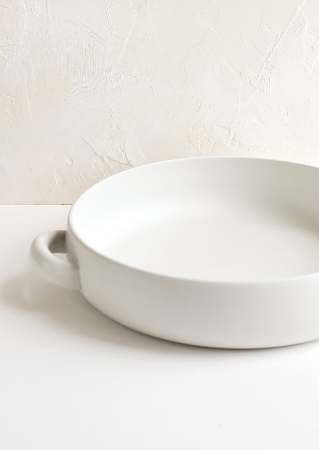 2: A round, shallow extra large serving bowl in white ceramic with curved handles at sides.