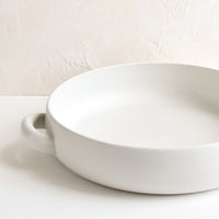 2: A round, shallow extra large serving bowl in white ceramic with curved handles at sides.