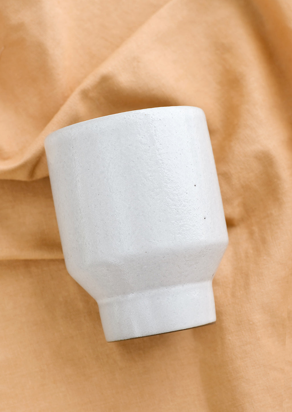 2: A white ceramic cup with a tapered base on brown fabric.