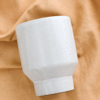 2: A white ceramic cup with a tapered base on brown fabric.