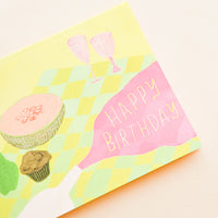 2: Greeting card with picnic setting, neon pink spilled wine puddle has letters reading "Happy Birthday"