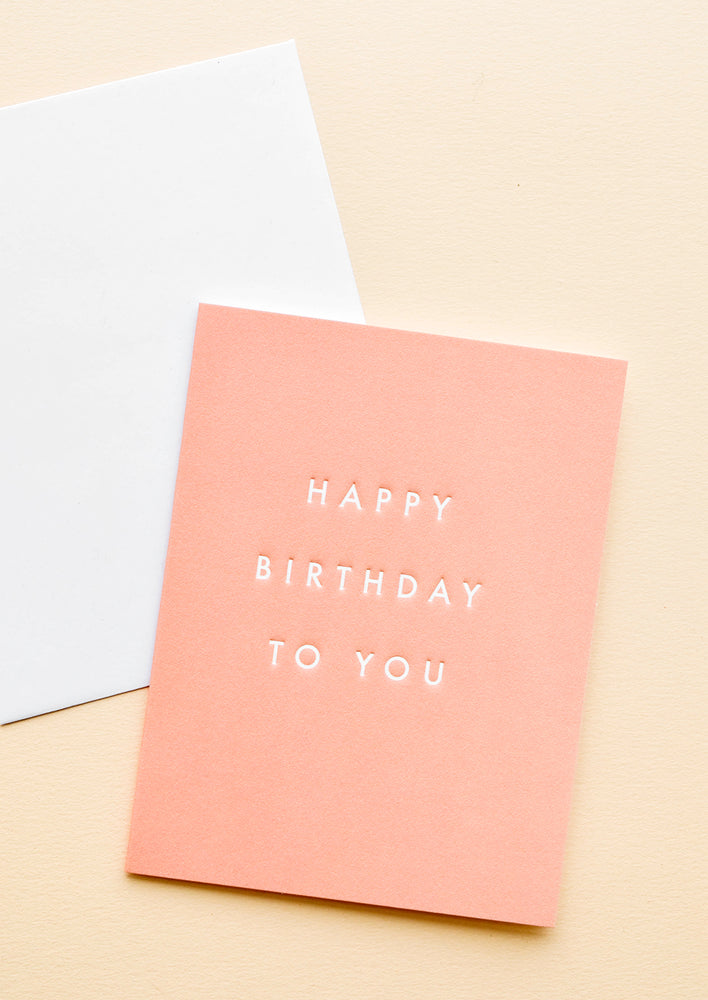 1: A white envelope and a coral colored greeting card with the phrase "happy birthday to you" in gold black letters.