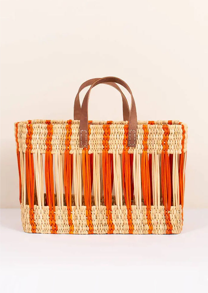 An oblong open weave basket in natural reed with bright orange stripes and brown leather handles.