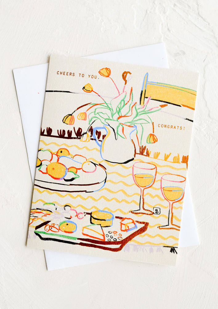 A greeting card with picnic illustration and copper lettering reading "Cheers to you. Congrats!"