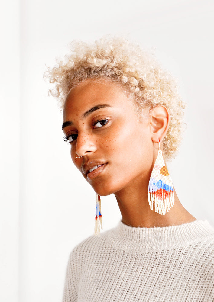 Model wears beaded fringe earrings and white top.