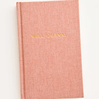 1: Fabric-covered journal in coral color with gold embossed text reading "The Well Journal"
