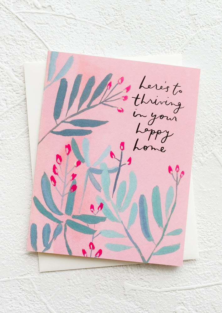 1: A pink greeting card with botanical background and text reading "Here's to thriving in your happy new home".