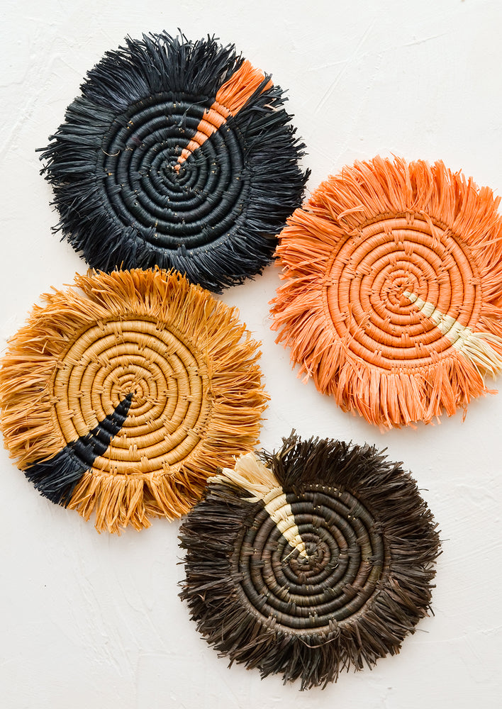 Four round raffia coasters in assorted earthy colors with fringed trim.
