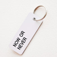 Now or Never: Small acrylic keychain, white background with black words that says "NOW OR NEVER"