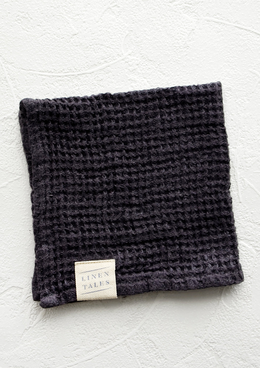 Charcoal: A folded waffle-weave linen washcloth in charcoal with a logo patch.