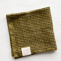 Olive: A folded waffle-weave linen washcloth in olive green with a logo patch.