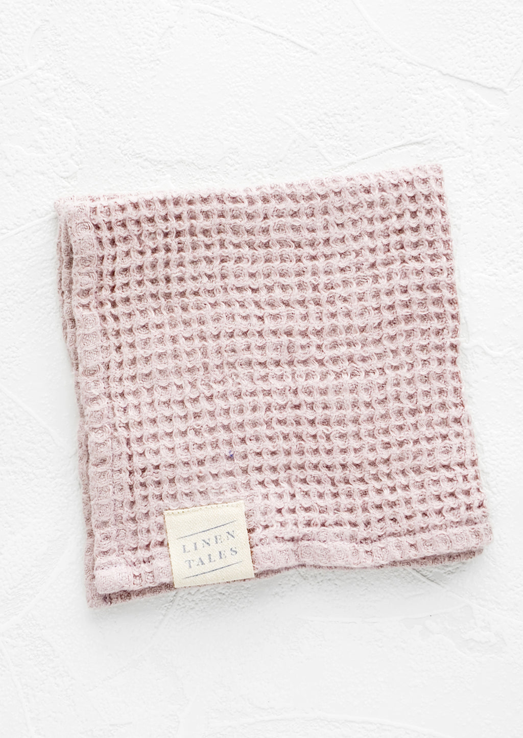 Rose: A folded waffle-weave linen washcloth in pale pink with a logo patch.