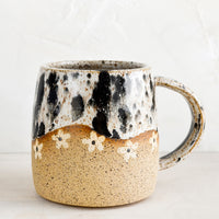 Appaloosa / Nude: A speckled ceramic coffee mug in black and white with wavy line of ivory daisies at middle.