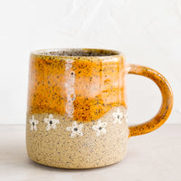 Calico / Nude: A speckled ceramic coffee mug in caramel with wavy line of ivory daisies at middle.