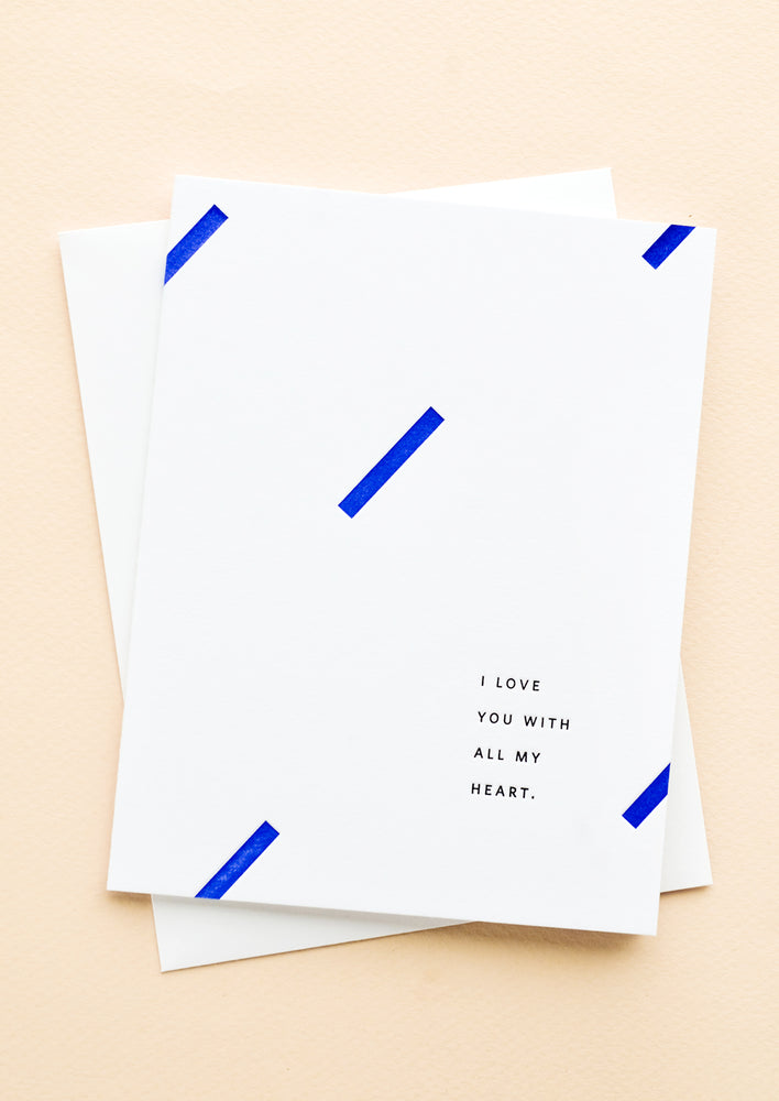 A white greeting card with blue geometric dashes and small caps text at corner reads "I love you with all my heart"